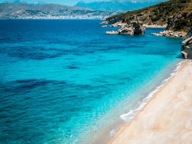 A bay with clear turquoise water, a pale strip of beach and hills and a town in the background