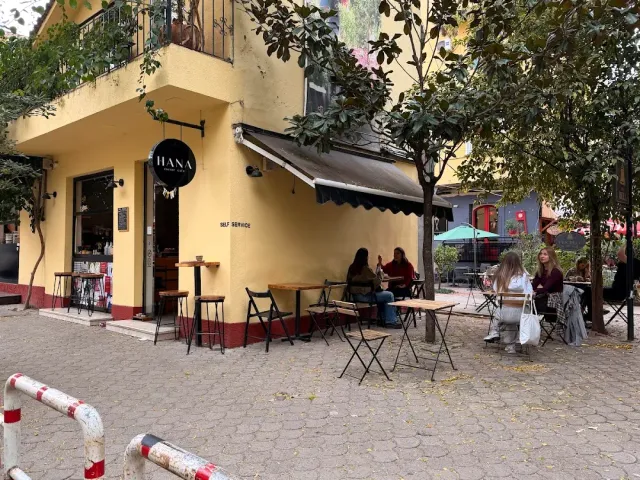 A yellow corner building with a small sign, tables outside and people sitting under the trees