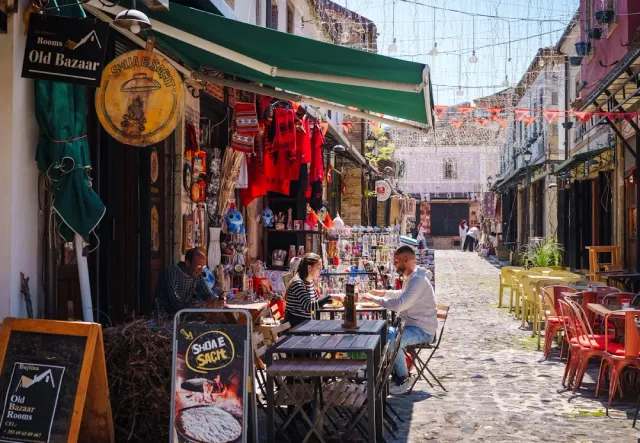 A stone bazaar alley with awnings, hanging souvenirs and diners around tables