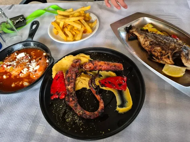 An octopus tentacle on a black plate with roasted peppers, beside fries and a whole fish