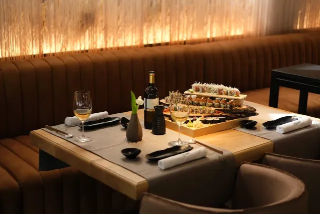 A table with a tiered sushi tray, a wine bottle and glasses, facing a padded brown bench