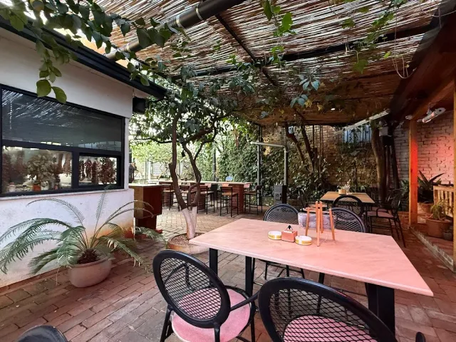 A garden courtyard covered with a reed roof, a pink table, black chairs and climbing greenery