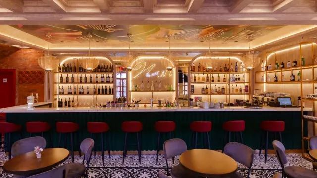 A long bar counter with red and grey stools and lit shelves of bottles behind