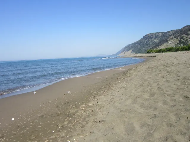 A long sandy beach with small waves, blue water and a green ridge at the end