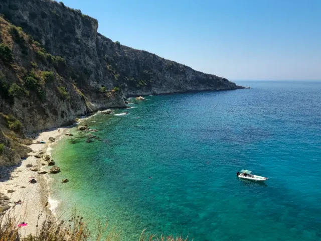 A high cliff dropping to turquoise water, a narrow pebble strip and a small anchored boat