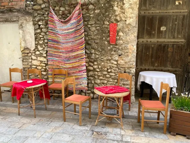 A colorful rag rug hanging on a stone wall, and before it small tables and wooden chairs
