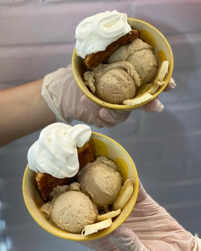Two gloved hands holding ice cream cups with whipped cream and white chocolate slices