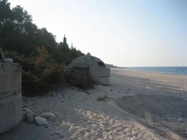 A round concrete bunker on the beach sand among pine trees, with the sea stretching to the right