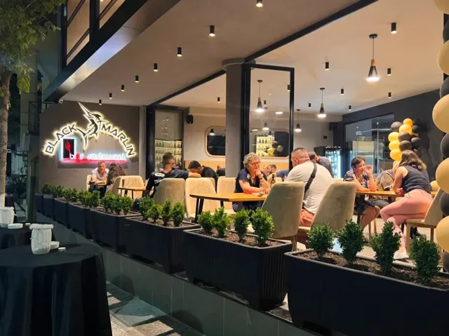 A terrace at night with a lit sign, diners at tables and planters of shrubs along the rail