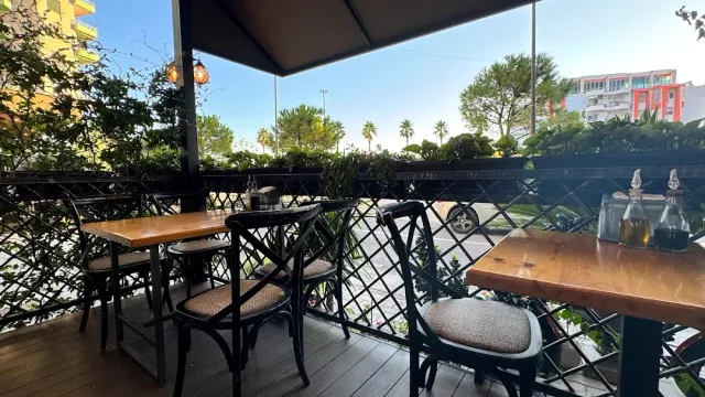 Wooden tables and dark chairs under an awning, behind a lattice railing with plants and palms