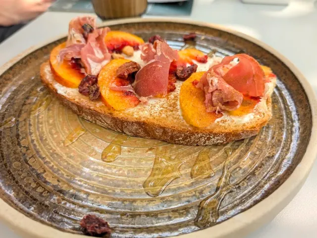 A slice of toasted bread with cheese spread, peach wedges and cured meat on a ceramic plate