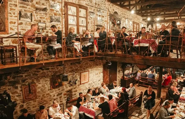 A two story stone hall with a wooden gallery, many diners and red striped cloths
