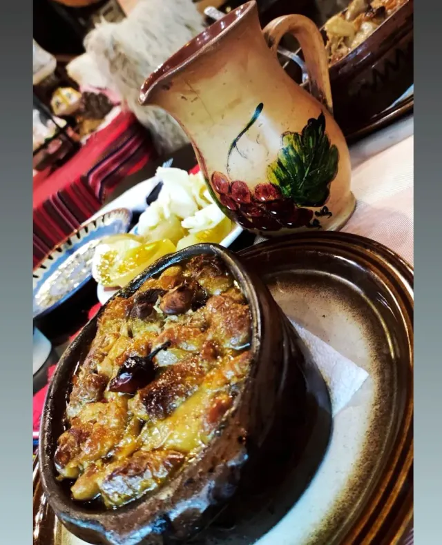 A clay pot with a golden baked dish, beside a painted jug and a plate of cheese and lemon