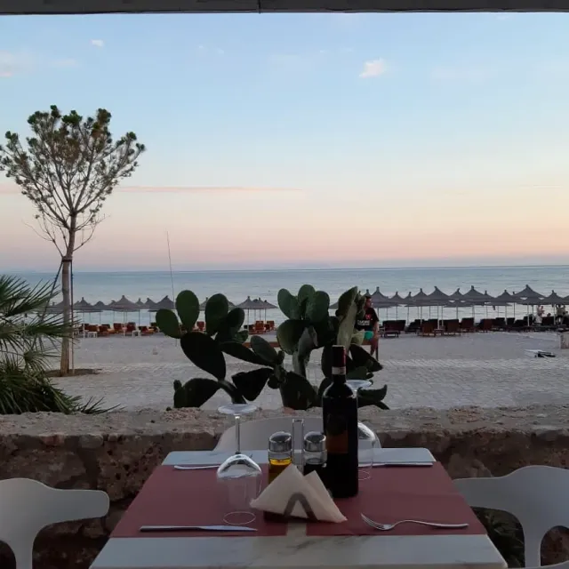A table with a pink cloth and a wine bottle, facing a beach with straw umbrellas and the sea at dusk