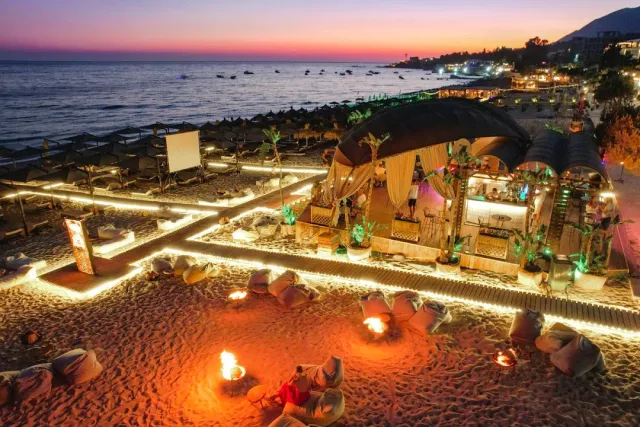 An aerial view at dusk of a lit beach venue with bean bags, fire bowls and a covered bar