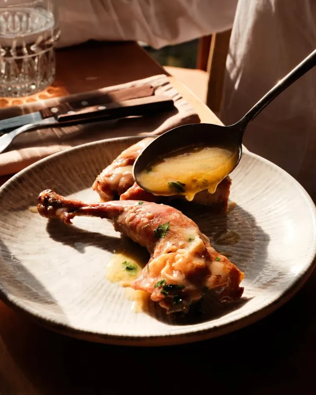 A spoon pouring yellow sauce over roasted legs on a ceramic plate, in warm sunlight