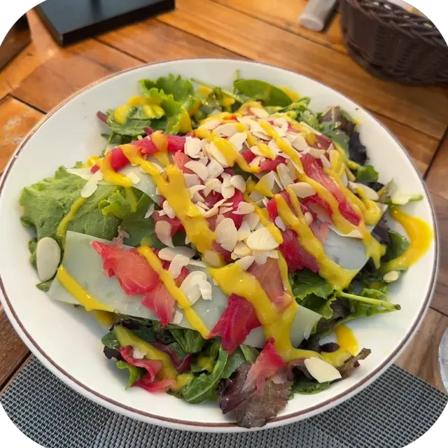 A white bowl with salad leaves, thin pink slices, sliced almonds and a yellow dressing
