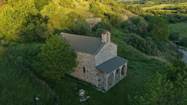 A small stone church with a tiled roof and an arcaded porch, surrounded by green slopes