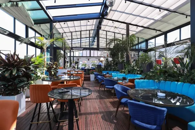 A glazed terrace with turquoise velvet benches, orange chairs and tropical plants