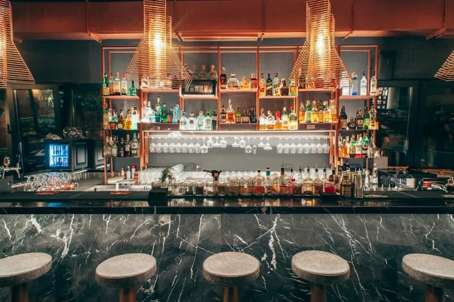 A dark marble counter with stools, behind it shelves of bottles and hanging straw lamps