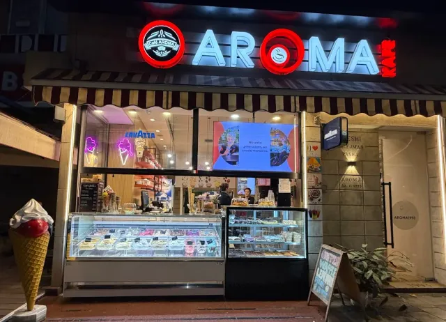 An ice cream shop lit at night with display cases, a neon sign and a giant cone at the entrance