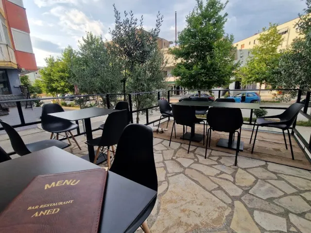 A paved terrace with black tables and chairs, potted olive trees and a menu in a cover