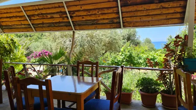A terrace roofed with wooden beams with a table and chairs, green vegetation and a strip of sea on the horizon