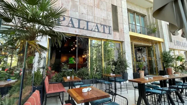 A street terrace with wooden tables, blue stools and dense greenery along a stone facade