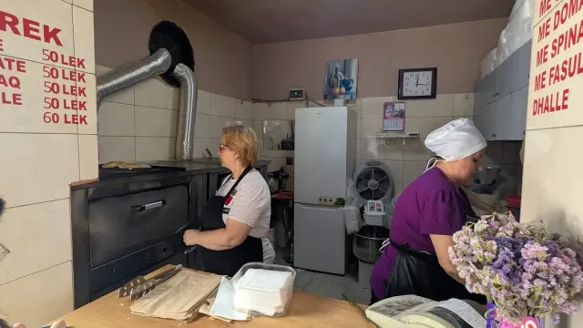 Two workers beside a metal oven in a bakery, a price list on the wall and trays on the counter