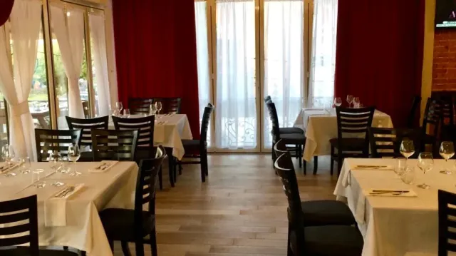 A dining room with white cloths, dark chairs and red curtains beside glass doors