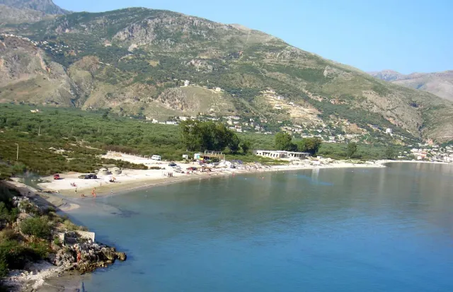 A calm bay with a white beach strip and a slope holding a village among olive trees
