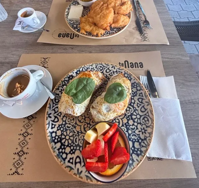 A decorated plate with fried eggs on toast and spinach leaves, a small fruit bowl and a cup of coffee