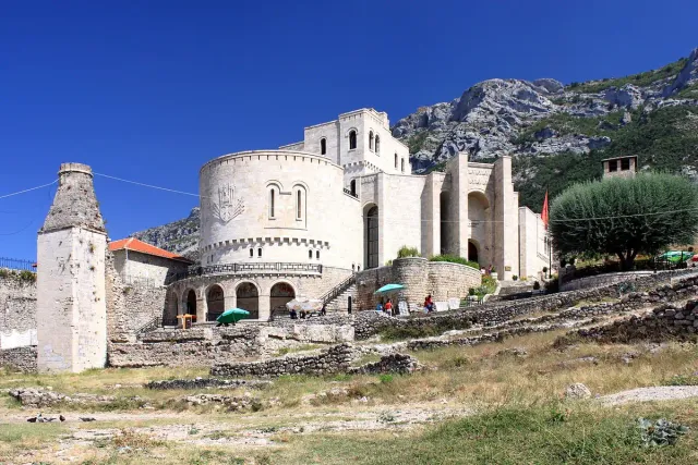 A white stone building with towers inside a castle compound, against a rocky mountain ridge
