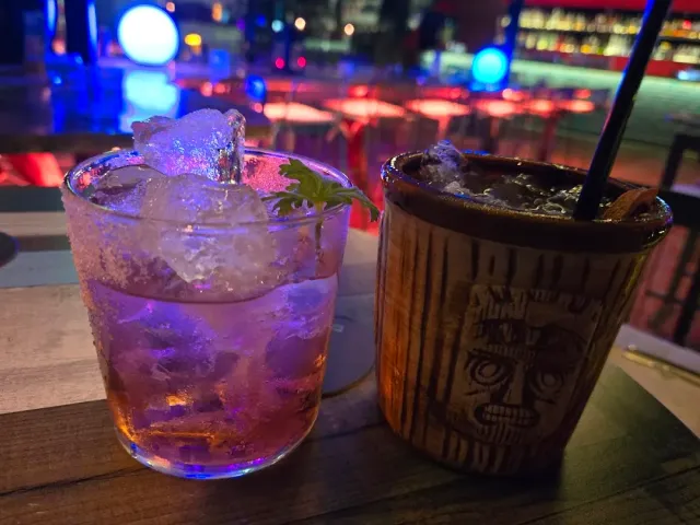 Two cocktails on a wooden table at night, one in a carved mug, against a backdrop of lights