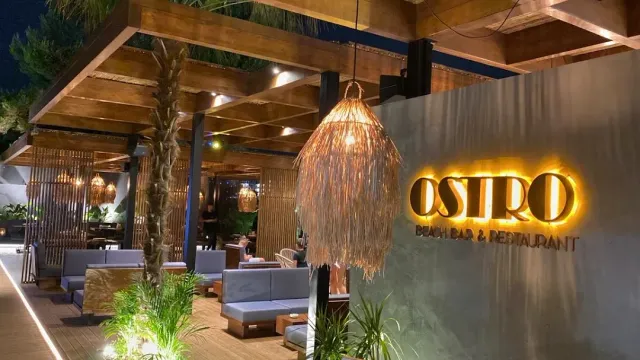 A wooden pergola at night with a large straw lamp, a lit sign on a concrete wall and sofas