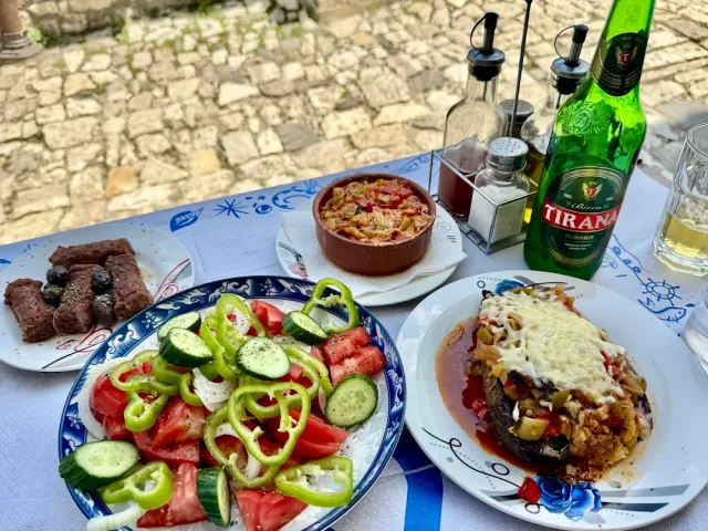 An outdoor table with a tomato and pepper salad, patties, baked eggplant and a beer bottle