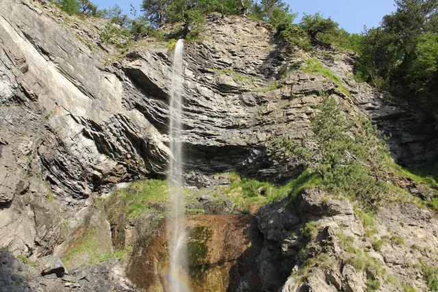 A tall narrow waterfall runs down a layered rock cliff among green trees