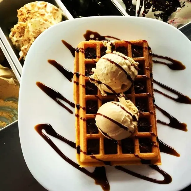 A Belgian waffle with two scoops of ice cream and chocolate sauce, next to ice cream tubs in the display