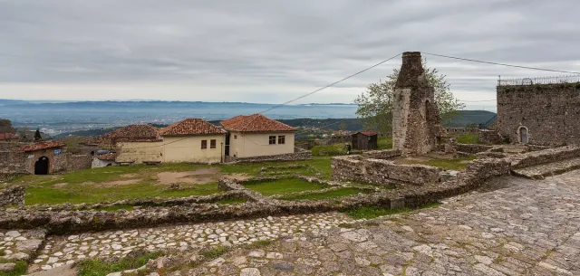 Ruined walls and a cobbled path inside a fortress, with a tower, houses and a view over the plain