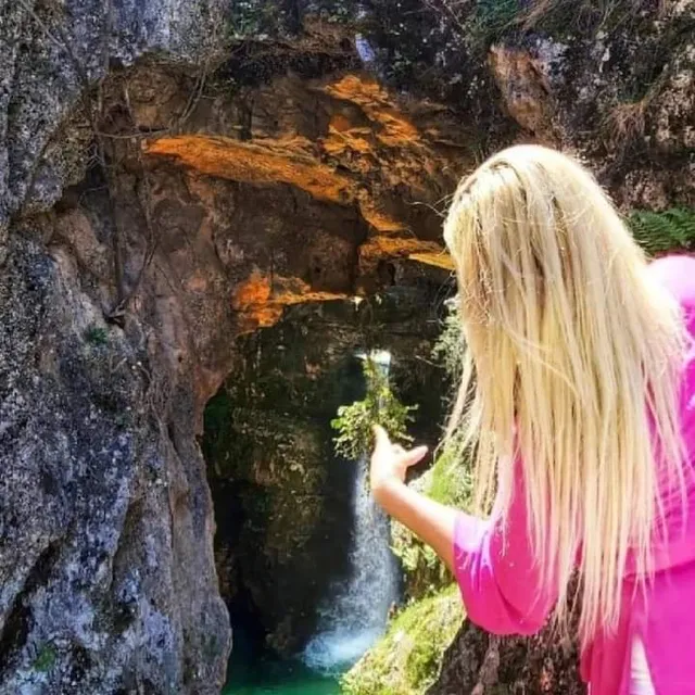 A thin waterfall drops into a greenish pool inside a rock gorge, as a hiker points at it