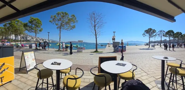 A view from under a canopy of round tables with yellow stools facing a promenade and a blue bay
