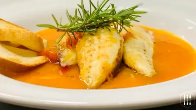Fish fillets in an orange sauce in a white bowl, with a rosemary sprig and toasted bread slices