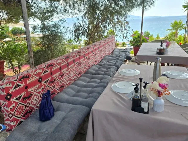 A long bench with grey cushions and a red patterned back, set tables and a sea view