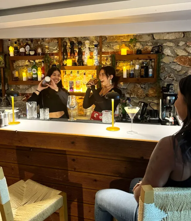 Two bartenders shaking cocktails behind a white counter, against a stone wall and shelves of bottles