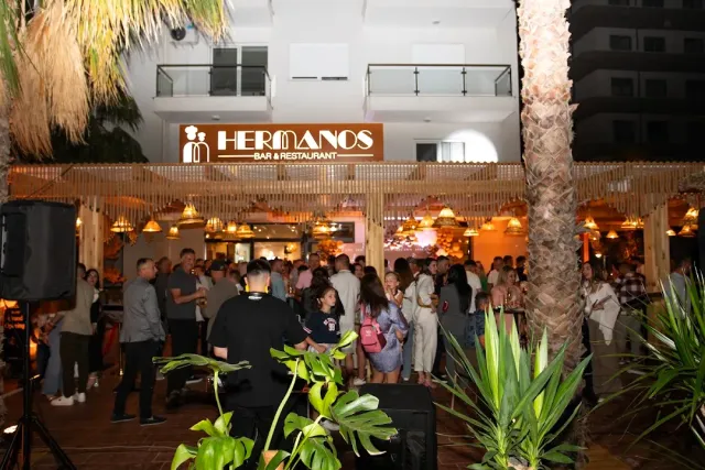 A dense crowd at night in front of a lit venue with a sign above, palm trees and a speaker at the side