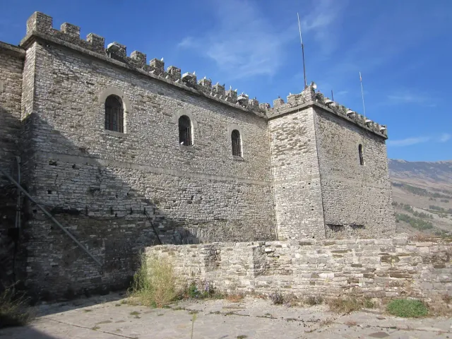 Grey stone castle ramparts with crenellations and pigeons along the edge, against a blue sky