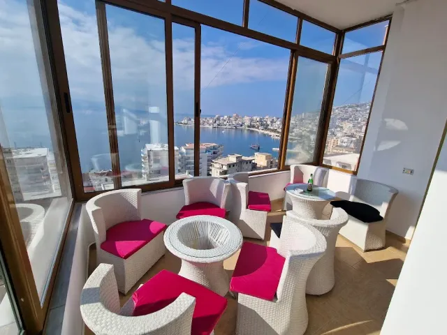 A glazed room with white rattan armchairs and pink cushions facing a bay and a town
