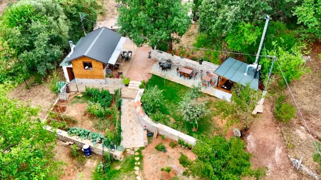 An aerial view of a wooden cabin with a seating terrace and a green garden among trees