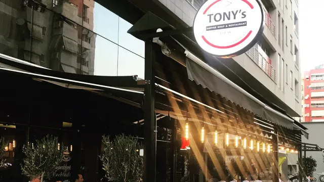 A round sign above a terrace awning, a string of lights and potted olive trees in the evening