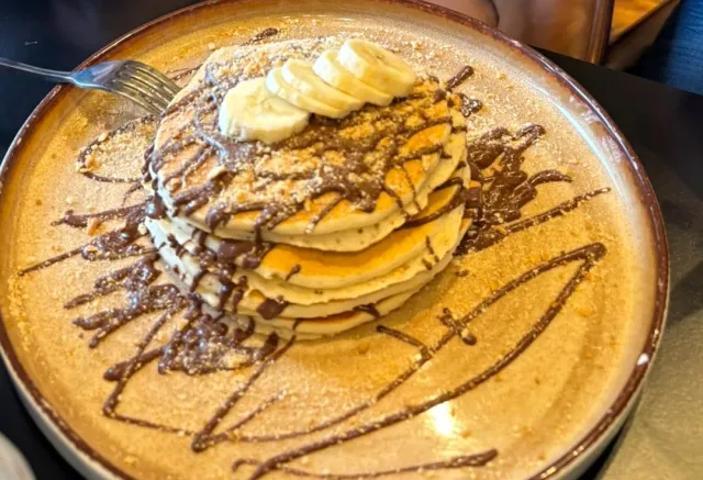 A stack of pancakes on a ceramic plate with banana slices and chocolate drizzle around